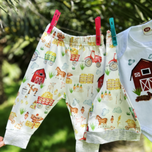 Handmade long-sleeve baby bodysuit with a farm-themed design, including a red barn, animals, and the text 'New to the Farm', displayed outdoors with green background